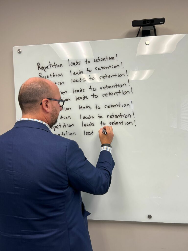 A photo of Greg writing "repetition leads to retention" on a whiteboard in repeating lines